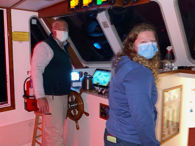 Two masked people in the wheelhouse of a boat